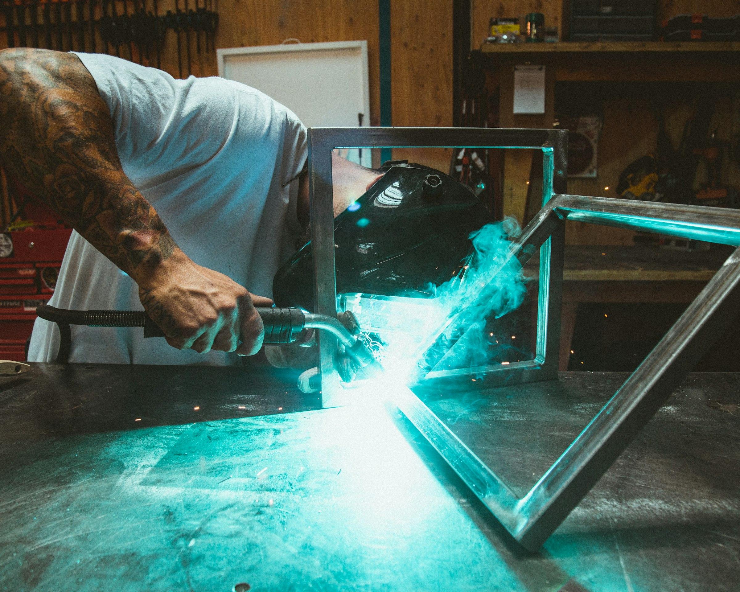 Welder joining a steel frame, the arc casting blue light across the workshop