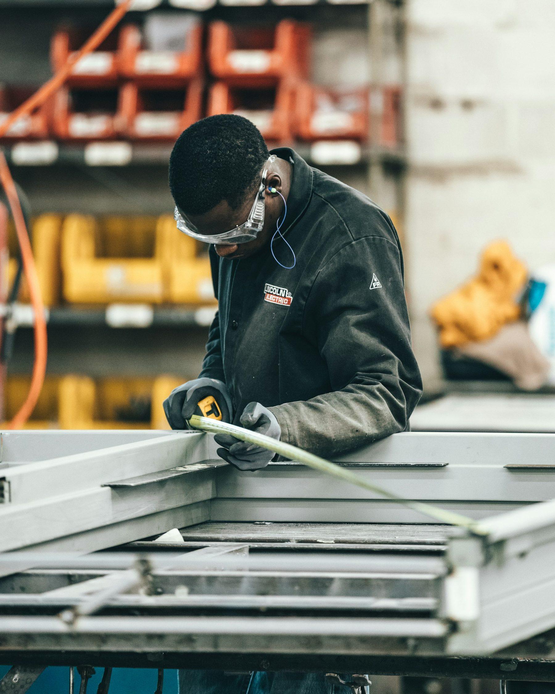 Technician in safety glasses measuring a fabricated metal frame in a workshop