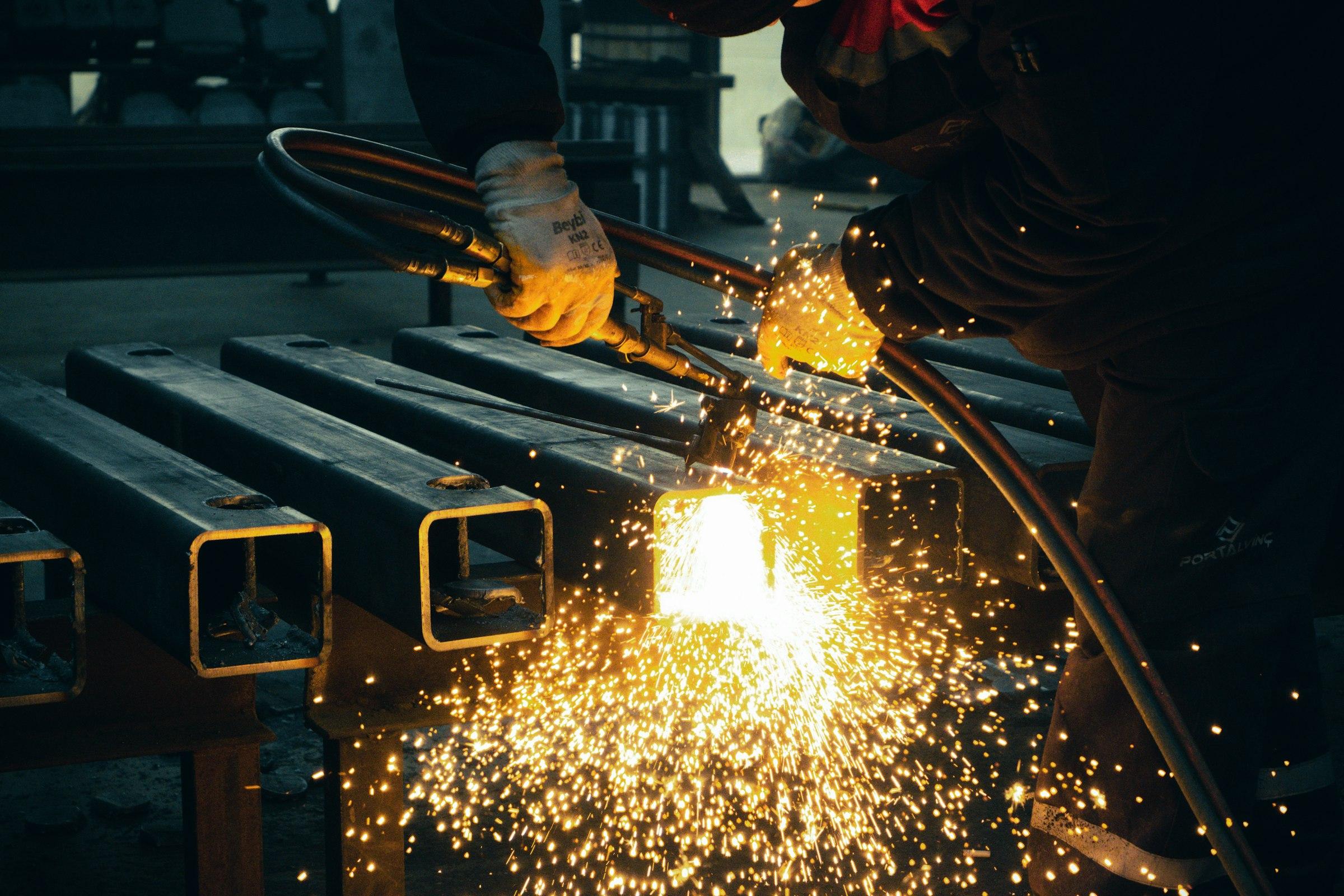 Fabricator welding rectangular steel sections, sparks spilling across the workbench